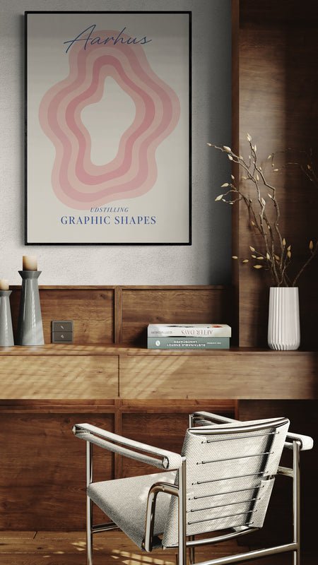 Plakat z napisem "Aarhus" i graficznym wzorem w odcieniach różu zawieszony nad drewnianym biurkiem w stylu retro z metalowym krzesłem i dekoracjami