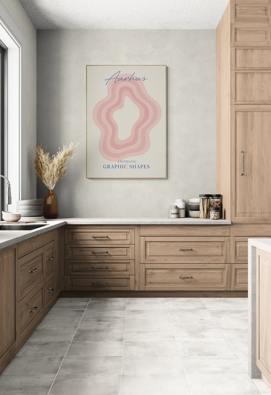 Plakat z napisem "Aarhus" i abstrakcyjnym wzorem w różowych odcieniach zawieszony nad blatem w jasnej kuchni z drewnianymi szafkami