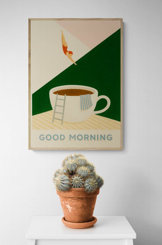 Plakat z napisem "Good Morning" i ilustracją filiżanki kawy, do której skacze postać, zawieszony na białej ścianie nad doniczką z kaktusem