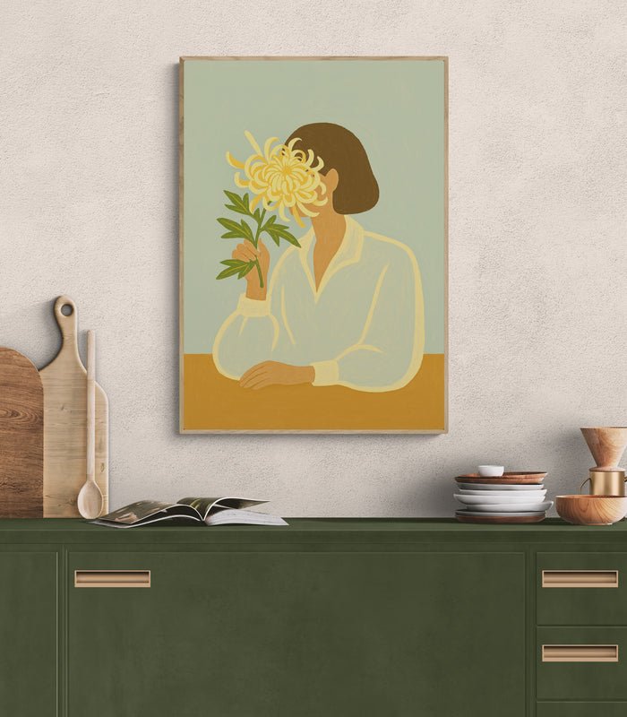 Plakat z ilustracją kobiety trzymającej chryzantemę, zawieszony nad zieloną kuchenną komodą z drewnianymi dodatkami i naczyniami