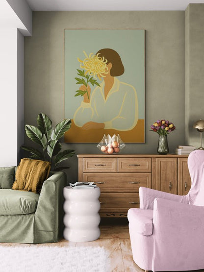 Plakat z ilustracją kobiety trzymającej chryzantemę, zawieszony nad drewnianą komodą w kolorowym salonie z różowym fotelem i zieloną sofą