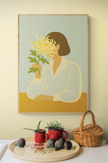Plakat z ilustracją kobiety trzymającej chryzantemę, zawieszony na pastelowej ścianie nad kuchennym blatem z czerwonymi kubkami i koszykiem
