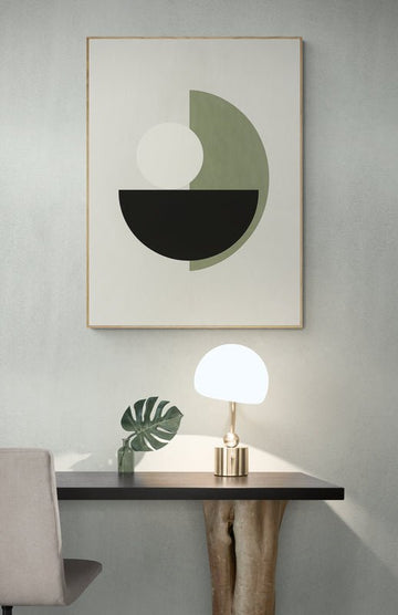 Plakat z geometrycznym motywem koła i półkola w zieleni, czerni i bieli zawieszony nad biurkiem z naturalną drewnianą nogą i lampką stołową