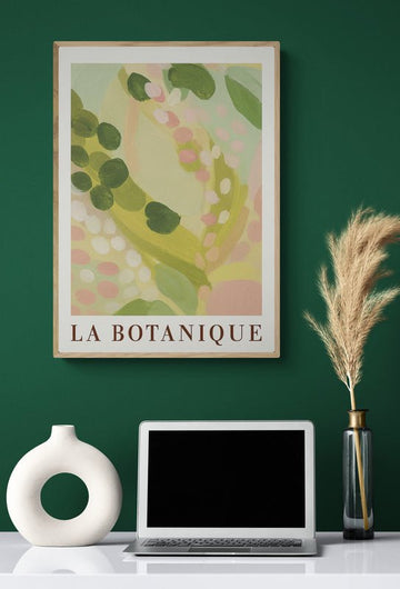 Plakat z napisem "La Botanique" i pastelowym motywem roślinnym zawieszony nad biurkiem z laptopem, dekoracyjnym wazonem i suszoną trawą na ciemnozielonej ścianie