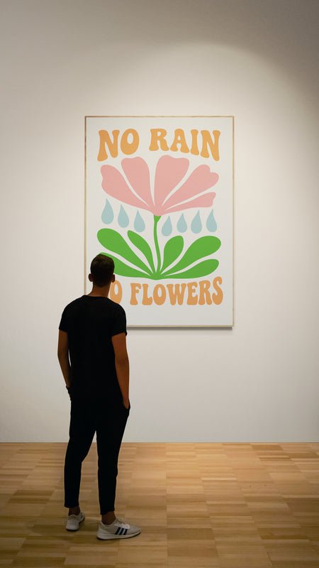 Plakat z napisem "No Rain No Flowers" i kolorowym kwiatem wystawiony na ścianie galerii, oglądany przez osobę stojącą na drewnianej podłodze